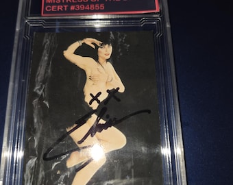 May include: A graded trading card featuring Elvira, Mistress of the Dark. The card has a black background with an image of Elvira. The card is encased in a clear protective holder. The text "ELVIRA MISTRESS OF THE DARK CERT #394855" is visible.