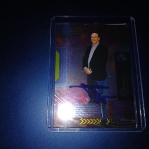 May include: A trading card encased in a clear plastic holder. The card features a man in a black blazer, blue jeans, and a white shirt. The card has the text "Jon Taffer" and "Mega Universe" printed on it. The card is signed in blue ink.