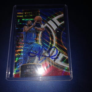 Puede incluir: Tarjeta de baloncesto coleccionable con un jugador con uniforme azul, en pleno salto, sosteniendo un balón. La tarjeta tiene un fondo holográfico y una firma con tinta azul. El nombre y el equipo del jugador están impresos en la parte inferior.