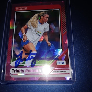 May include: A Trinity Rodman football trading card. The card features a photo of Rodman in a white jersey with the number 22. The card has a red and silver background and includes the FIFA logo and the words "United States."