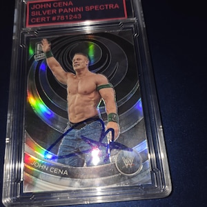 Op de afbeelding: Een John Cena Silver Panini Spectra verzamelkaart, in een beschermhoes. De kaart toont John Cena, ontbloot bovenlijf, met zijn handtekening. Tekst op de kaart luidt "JOHN CENA", "SILVER PANINI SPECTRA" en "CERT #781243".