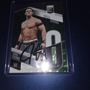 Op de afbeelding: Een John Cena-verzamelkaart in een beschermhoesje. De kaart toont een foto van John Cena, de tekst "SPELL BOUND" en zijn handtekening. De kaart heeft een groen-witte achtergrond met het nummer "0".