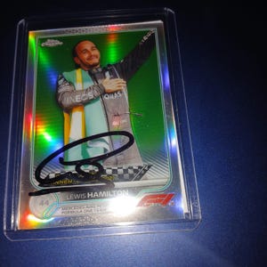 May include: A collectible trading card featuring Lewis Hamilton, with a holographic green background. The card has an autograph and the text "WINNER" and "2021". The card is in a protective case. The card also has the text "LEWIS HAMILTON" and "MERCEDES-AMG PETRONAS FORMULA ONE TEAM".