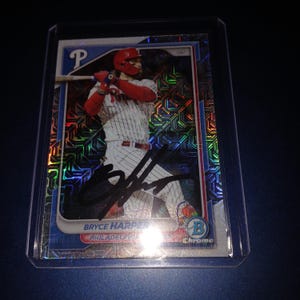 Puede incluir: Una tarjeta de béisbol con Bryce Harper de los Filis de Filadelfia. La tarjeta tiene un fondo holográfico con un borde azul. Harper está en posición de bateo, con un casco rojo y uniforme de los Filis. La tarjeta está en una funda protectora.