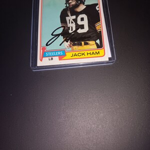 Op de afbeelding: Een vintage Topps-verzamelkaart met Jack Ham, een linebacker van de Pittsburgh Steelers. De kaart is in goede staat en toont Ham in zijn uniform met nummer 59 op zijn shirt. De kaart heeft een blauwe achtergrond en de tekst "STEELERS LB" is in wit onderaan gedrukt.