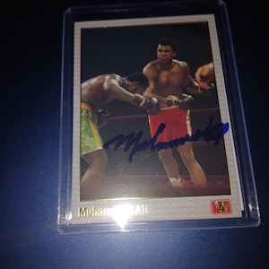May include: A trading card featuring a boxing match between two athletes. One boxer is wearing red shorts and the other is wearing green and yellow shorts. The card is signed with a blue pen. The card reads "Muhammad Ali".