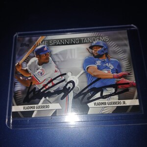 May include: A baseball card featuring Vladimir Guerrero and Vladimir Guerrero Jr. The card has the text "TIME SPANNING TANDEMS" at the top. Both players are in their baseball uniforms. The card is encased in a protective sleeve.