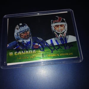 May include: A hockey trading card showing two goalies wearing helmets. The card has the text "O CANADA" and the names "PATRICK ROY" and "MARTIN BRODEUR". The card is in a clear plastic holder.