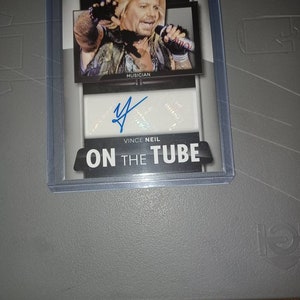 May include: A trading card featuring a black and white photo of Vince Neil, a musician, with the text "Panini Americana" and "On the Tube" on the card. The card has a silver background and a white signature area with a blue signature.