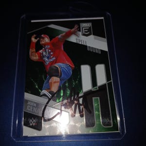 Puede incluir: Una tarjeta coleccionable Elite "Spell Bound" de John Cena. La tarjeta presenta a Cena con una camisa roja y pantalones cortos azules, con su firma en rotulador negro. La tarjeta está dentro de una funda protectora de plástico.