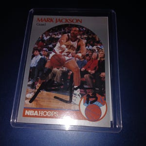 May include: A Mark Jackson NBA Hoops basketball card featuring a player in action, wearing a New York Knicks uniform. The card is encased in a protective sleeve. The card has the text "MARK JACKSON" and "Guard".