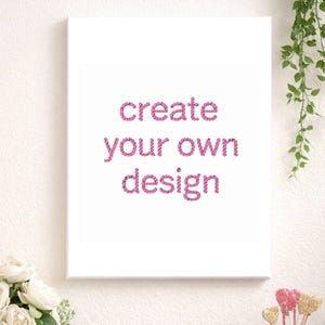 May include: A white canvas with the text "create your own design" in pink glitter lettering. The canvas is displayed on a white wall, with decorative elements including flowers and a candle.