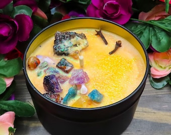 May include: A yellow candle with embedded crystals and spices in a black tin container. The candle is surrounded by pink and purple flowers.