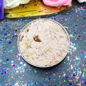 May include: A close-up of a small, round container filled with a light brown mixture. The container is sitting on a sparkly, iridescent surface with a blue background.