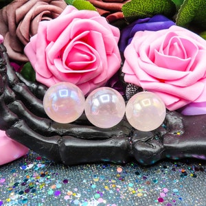 A black skeletal hand with glittery, pink painted nails holding three pink angel aura rose quartz spheres of varying sizes.