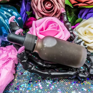 May include: A black skeletal hand holding a dark glass bottle with a dropper. The bottle is surrounded by artificial flowers in shades of pink, purple, and white.