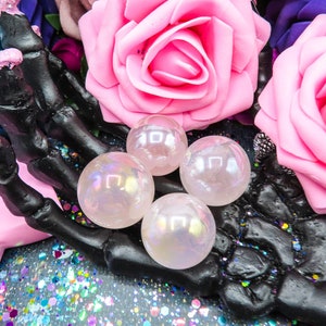 Four iridescent, pink, angel aura rose quartz spheres rest on a black skeletal hand. The spheres are translucent and display a subtle rainbow effect. They are surrounded by artificial pink roses and a dark background with iridescent glitter.