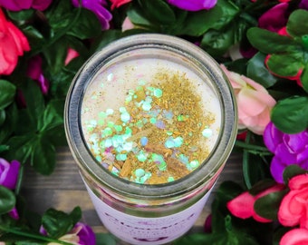 May include: A clear glass jar candle with a white wax interior, adorned with gold and iridescent glitter. The candle is surrounded by a vibrant arrangement of pink, red, and purple flowers and green leaves, creating a colorful floral display.