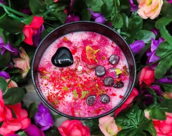 May include: A black candle jar filled with a pink and red wax blend. The wax is studded with small black and purple crystals and dried flower petals. The jar is surrounded by pink and purple flowers.