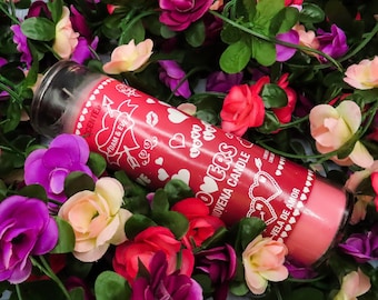 May include: A red scented Lovers Novena candle with white text and heart designs, surrounded by colorful artificial flowers. The candle has a pink base and is approximately 8 inches tall.