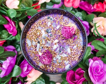 May include: A purple and gold glitter candle with dried flowers and crystals embedded in the wax. The candle is surrounded by pink and purple artificial flowers.