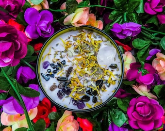 May include: A purple, white and yellow candle with crystals and dried herbs embedded in the wax. The candle is surrounded by artificial flowers in shades of pink, purple and white.