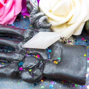 K&ouml;nnte beinhalten: Ein wei&szlig;er Kristall-Anh&auml;nger in Form eines Diamanten, der an einer silbernen Kette h&auml;ngt. Der Anh&auml;nger liegt auf einer schwarzen Skeletthand, umgeben von buntem Glitzer.