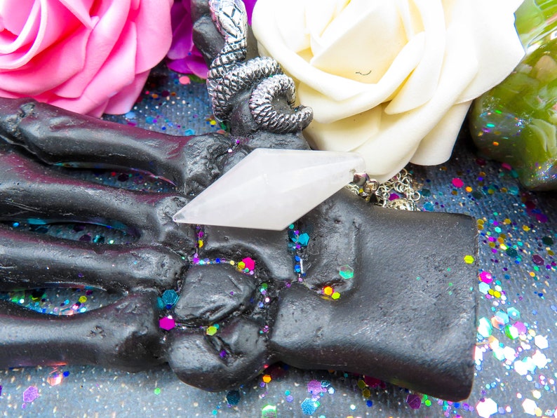 K&ouml;nnte beinhalten: Ein wei&szlig;er Kristall-Anh&auml;nger in Form eines Diamanten, der an einer silbernen Kette h&auml;ngt. Der Anh&auml;nger liegt auf einer schwarzen Skeletthand, umgeben von buntem Glitzer.