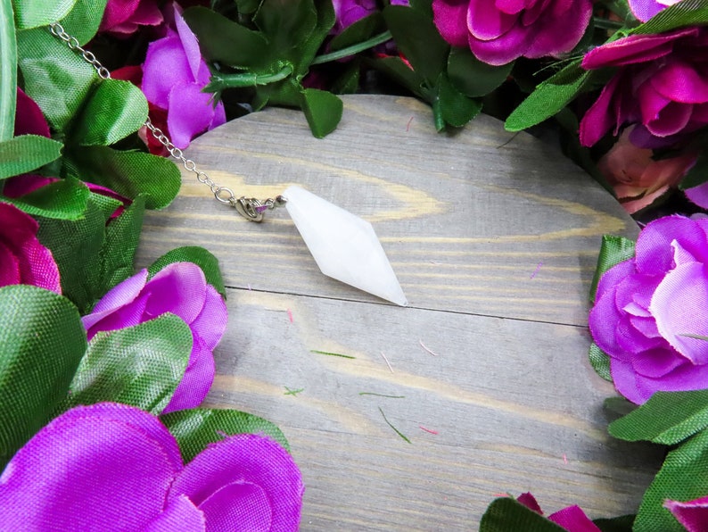 K&ouml;nnte beinhalten: Wei&szlig;er Kristallpendel an einer silbernen Kette, der auf einer Holzoberfl&auml;che liegt, umgeben von lila Blumen.
