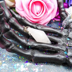 K&ouml;nnte beinhalten: Ein wei&szlig;er Kristall-Anh&auml;nger in Form eines Diamanten, mit einer silbernen Kette, der in einer schwarzen Skeletthand ruht.
