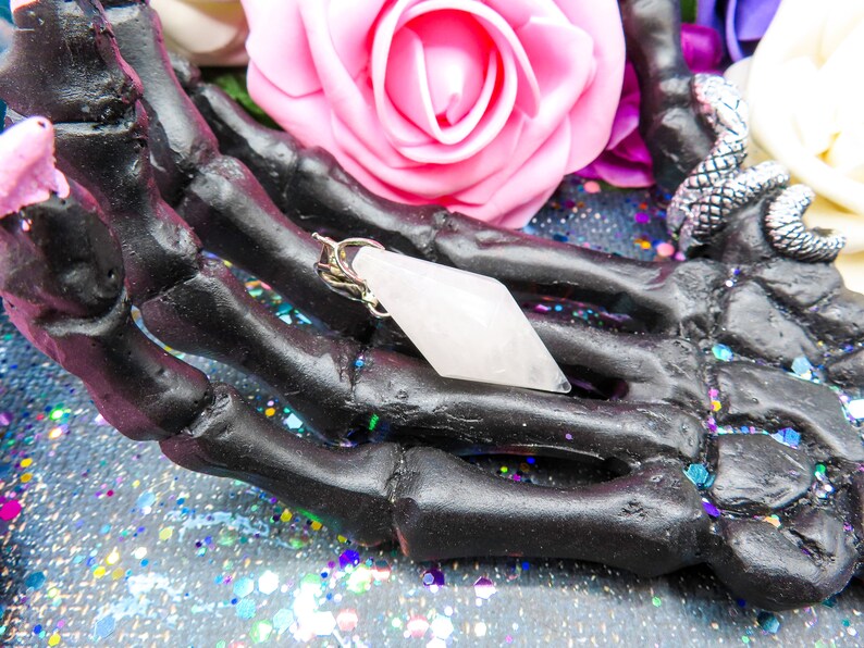 K&ouml;nnte beinhalten: Ein wei&szlig;er Kristall-Anh&auml;nger in Form eines Diamanten, mit einer silbernen Kette, der in einer schwarzen Skeletthand ruht.