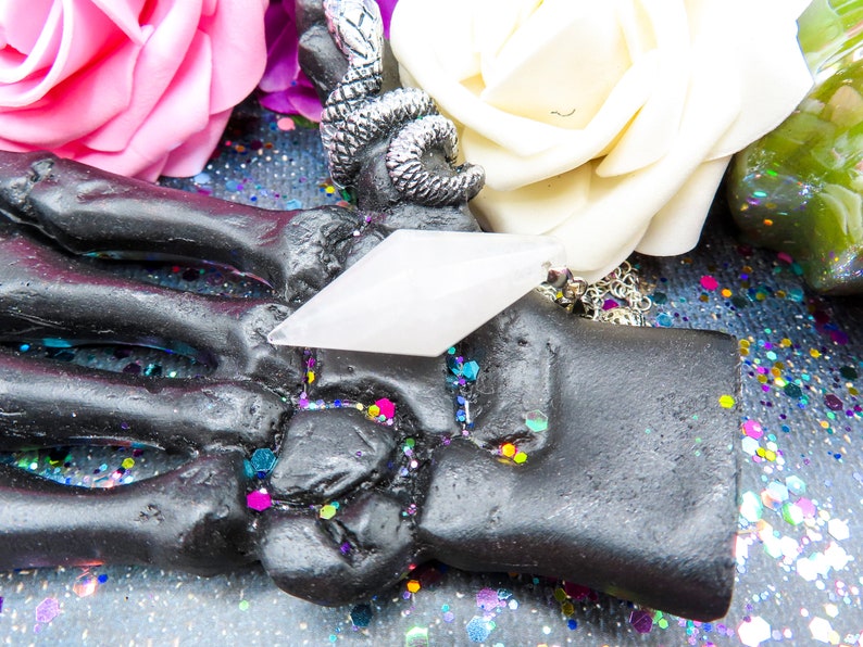 K&ouml;nnte beinhalten: Ein wei&szlig;er Kristall-Anh&auml;nger in Form eines Diamanten, der an einer silbernen Kette h&auml;ngt. Der Anh&auml;nger liegt auf einer schwarzen Skeletthand, umgeben von buntem Glitzer.