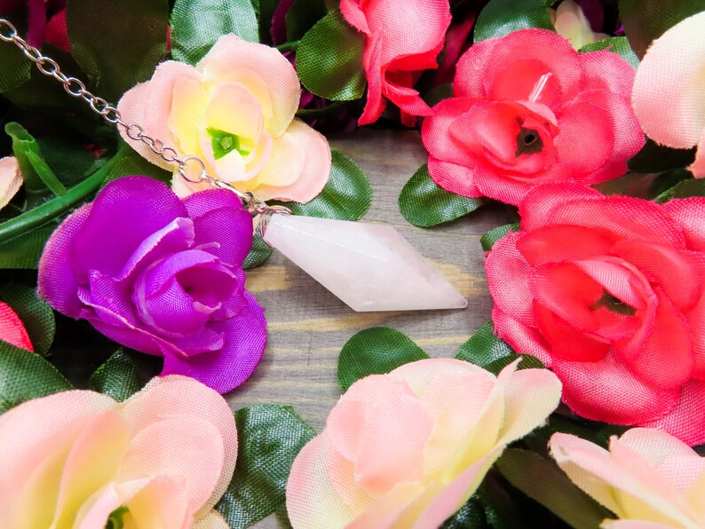 K&ouml;nnte beinhalten: Ein wei&szlig;er Kristallpendel mit einer silbernen Kette, umgeben von k&uuml;nstlichen rosa, wei&szlig;en und violetten Blumen.
