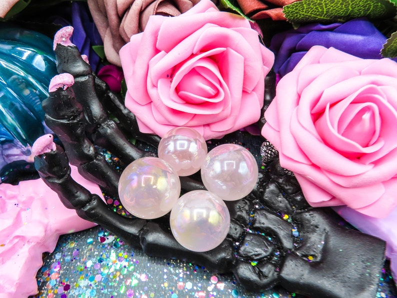 Four iridescent, pink, angel aura rose quartz spheres rest on a black skeletal hand. The spheres are translucent and display a subtle rainbow effect. They are surrounded by artificial pink roses and a dark background with iridescent glitter.