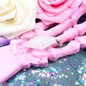 K&ouml;nnte beinhalten: Ein wei&szlig;er Kristall-Anh&auml;nger in Form eines Diamanten h&auml;ngt an einer silbernen Kette. Der Anh&auml;nger liegt auf einem rosa handf&ouml;rmigen Objekt, umgeben von buntem Glitzer.