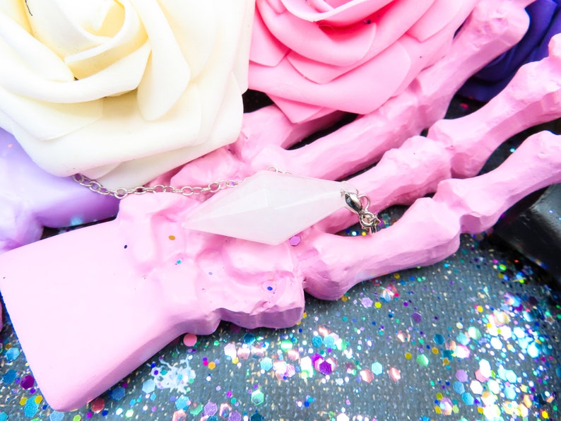 K&ouml;nnte beinhalten: Ein wei&szlig;er Kristall-Anh&auml;nger in Form eines Diamanten h&auml;ngt an einer silbernen Kette. Der Anh&auml;nger liegt auf einem rosa handf&ouml;rmigen Objekt, umgeben von buntem Glitzer.