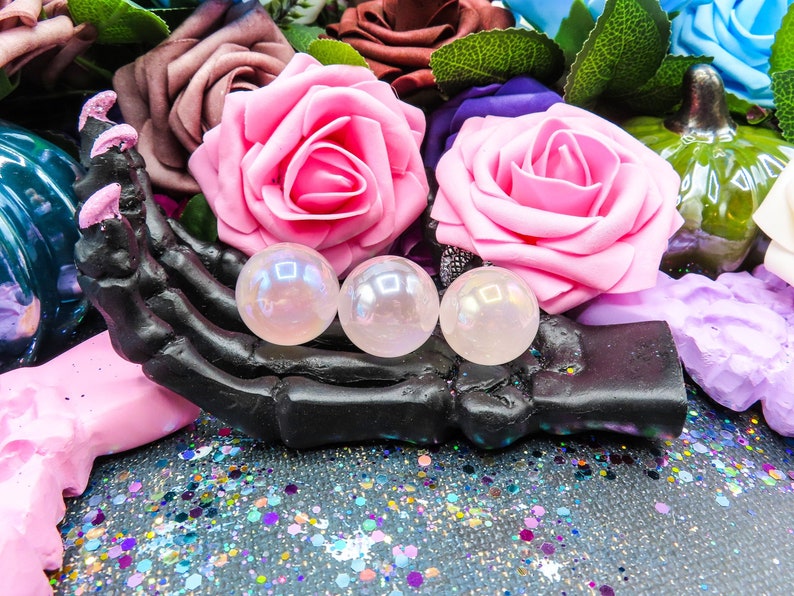 A black skeletal hand with glittery, pink painted nails holding three pink angel aura rose quartz spheres of varying sizes.