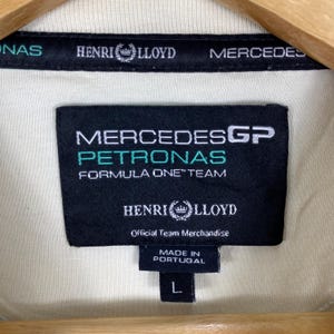May include: A cream-coloured garment featuring a black interior neck label with white and teal text reading Mercedes GP Petronas Formula One Team, Henri Lloyd, and Official Team Merchandise, with a small tag below indicating Made in Portugal and size L.