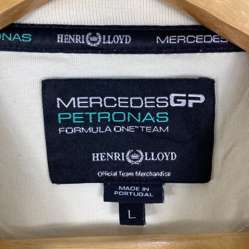 May include: A cream-coloured garment featuring a black interior neck label with white and teal text reading Mercedes GP Petronas Formula One Team, Henri Lloyd, and Official Team Merchandise, with a small tag below indicating Made in Portugal and size L.