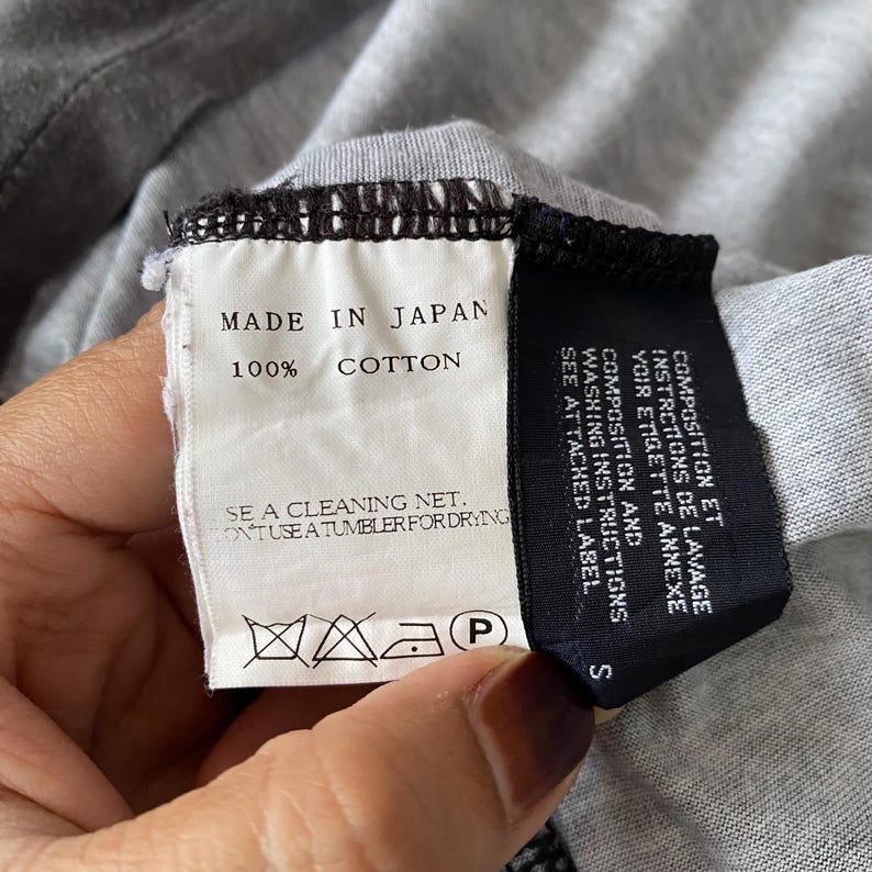 Puede incluir: Una etiqueta de cuidado de prenda de tela blanca con texto negro que dice MADE IN JAPAN y 100% COTTON, junto a una etiqueta negra con instrucciones de lavado en franc&eacute;s e ingl&eacute;s. La etiqueta blanca incluye s&iacute;mbolos de limpieza y secado.