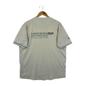 May include: A beige short-sleeved t-shirt featuring the Mercedes GP Petronas Formula One Team logo printed in black and teal on the back. The shirt has a crew neckline and thin teal piping along the edges of the sleeves, displayed on a wooden hanger.