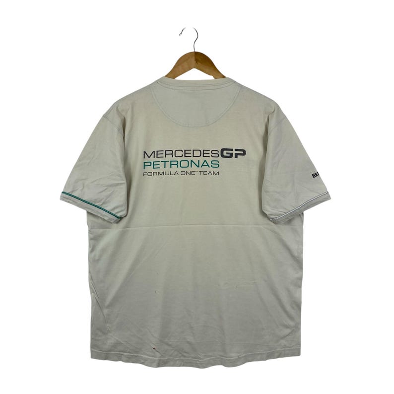 May include: A beige short-sleeved t-shirt featuring the Mercedes GP Petronas Formula One Team logo printed in black and teal on the back. The shirt has a crew neckline and thin teal piping along the edges of the sleeves, displayed on a wooden hanger.