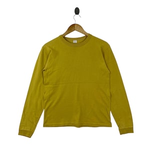 May include: Mustard yellow long-sleeve shirt. The shirt has a crew neck and a relaxed fit. The garment is hanging on a wooden hanger. The shirt is made of a soft, comfortable material.