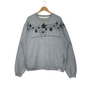 Puede incluir: Una sudadera gris con cuello redondo y mangas largas. La sudadera presenta un diseño floral negro y el texto "Welcome to The Imagination Paradise of NARCOTIC 2003". La prenda está colgada de una percha de madera.