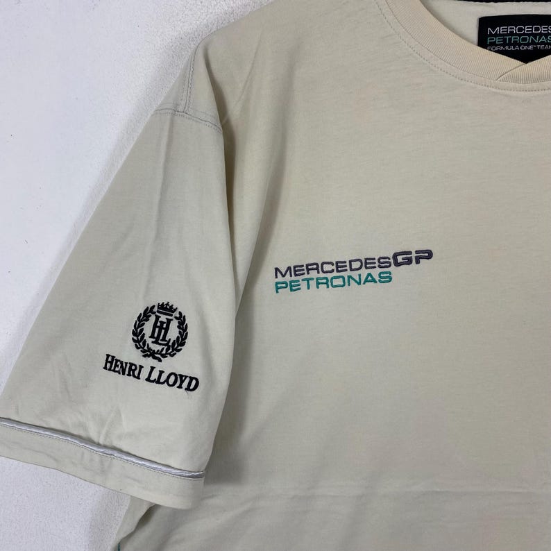 May include: A cream-coloured short-sleeve t-shirt featuring the Mercedes GP Petronas logo in dark blue and teal on the chest. The right sleeve displays a black embroidered Henri Lloyd logo with a crown and laurel wreath design.