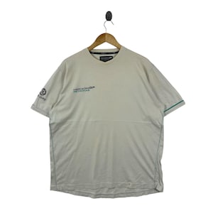 May include: A beige short-sleeve t-shirt featuring the Mercedes GP Petronas logo on the chest and the Lloyd logo on the right sleeve. The shirt has a crew neckline and thin teal piping accents along the sleeves and side seams of the garment.
