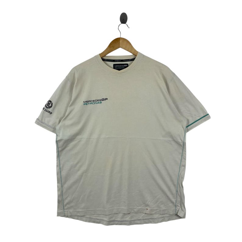 May include: A beige short-sleeve t-shirt featuring the Mercedes GP Petronas logo on the chest and the Lloyd logo on the right sleeve. The shirt has a crew neckline and thin teal piping accents along the sleeves and side seams of the garment.
