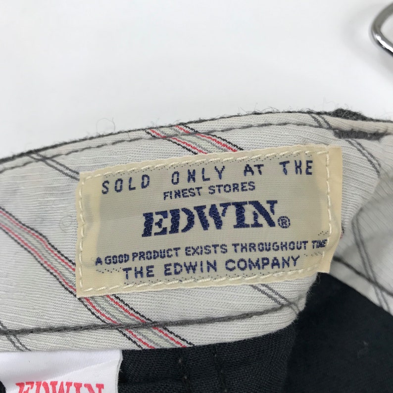 K&ouml;nnte beinhalten: Nahaufnahme eines Kleidungsetiketts mit dem Markennamen "EDWIN" in Marineblau. Das Etikett ist beige mit marineblauem Text, der "SOLD ONLY AT THE FINEST STORES" und "A GOOD PRODUCT EXISTS THROUGHOUT THE THE EDWIN COMPANY" lautet.