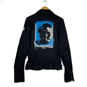 Könnte beinhalten: Schwarze Jacke mit langen Ärmeln und einem blauen und weißen Grafikdruck eines Gesichts auf der Rückseite. Die Grafik befindet sich in einem quadratischen Rahmen mit weißem Rand. Die Jacke hat einen Reißverschluss und zwei Reißverschlusstaschen an den Seiten.