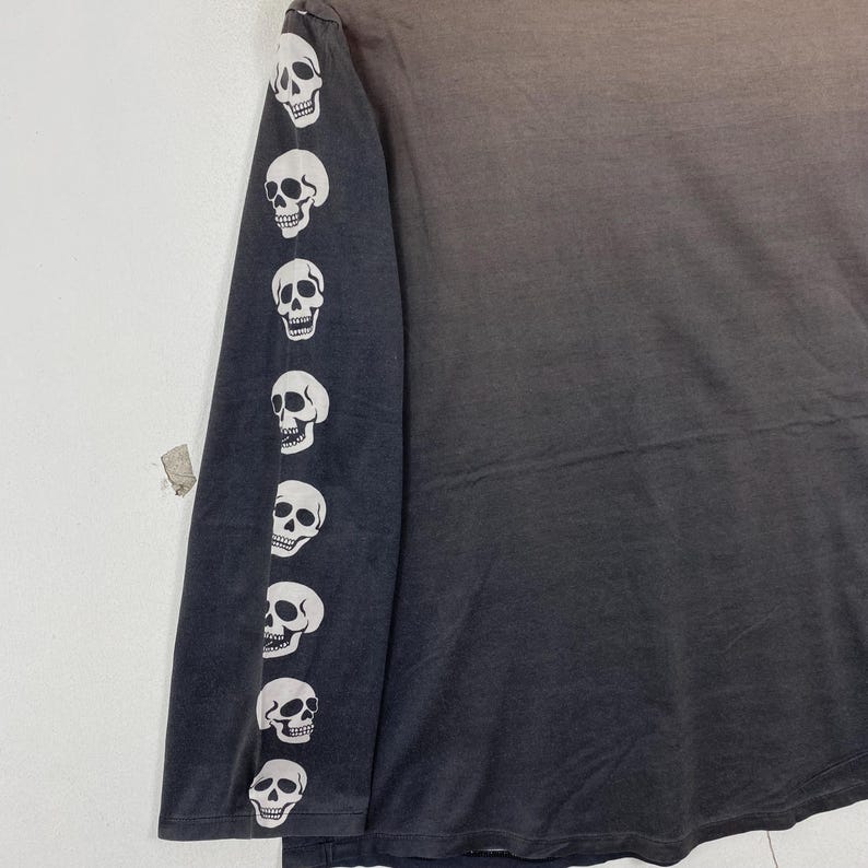 Puede incluir: Una camiseta de manga larga con un degradado de color gris oscuro a gris claro. La manga izquierda est&aacute; adornada con una fila vertical de ocho gr&aacute;ficos de calaveras blancas impresos en un patr&oacute;n repetitivo desde el hombro hasta el pu&ntilde;o.